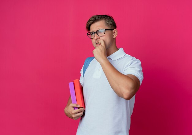 Free photo looking at side concerned young handsome male student wearing back bag and glasses holding books and bites nails isolated on pink wall