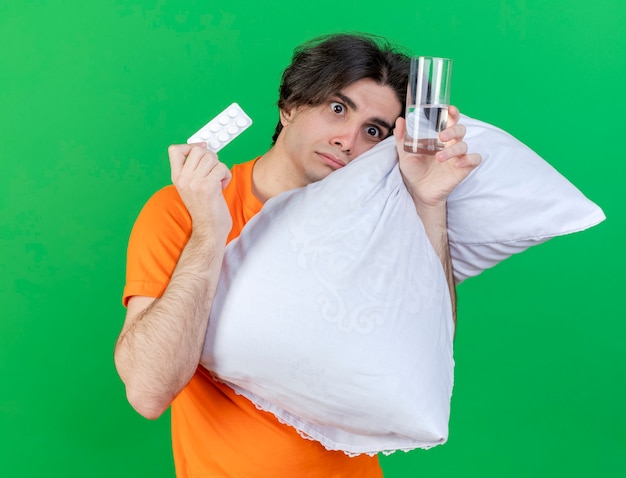 Foto gratuita guardando il lato interessato giovane uomo malato abbracciò il cuscino e tenendo il bicchiere d'acqua con le pillole isolato su sfondo verde