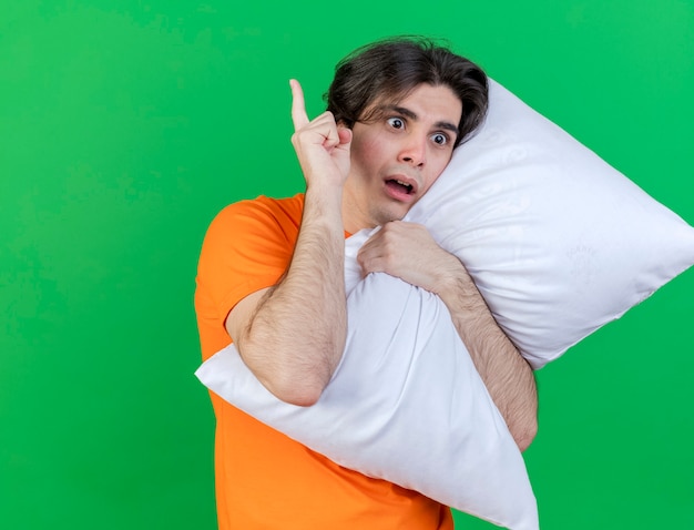 Free photo looking at side concerned young ill man hugged pillow and points at up isolated on green background