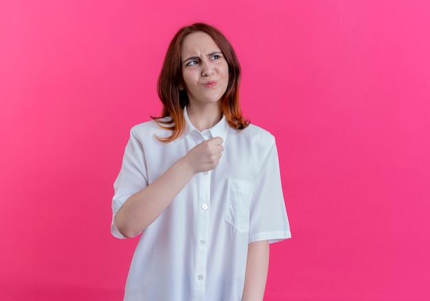 Looking at side concerned young redhead girl holding fist isolated on pink background