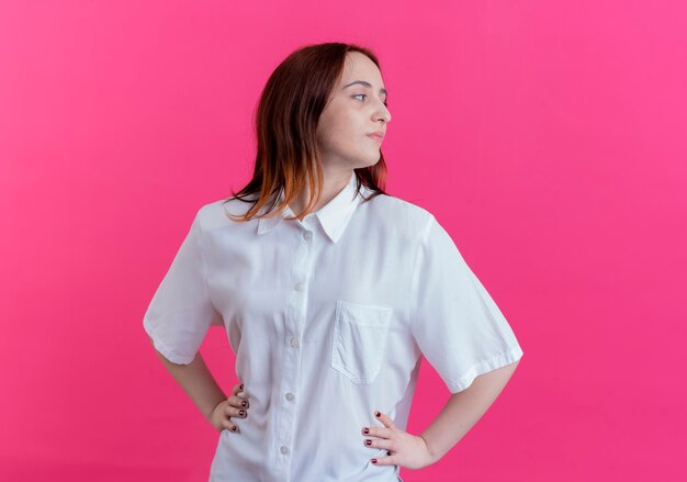 Free photo looking at side confident young redhead girl putting hands on hip isolated on pink background