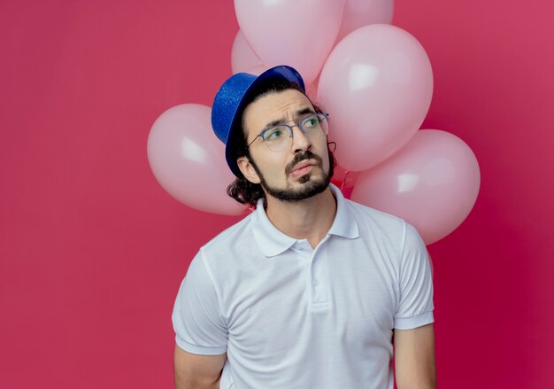 Looking at side confused handsome man wearing glasses and blue hat standing in front balloons isolated on pink wall