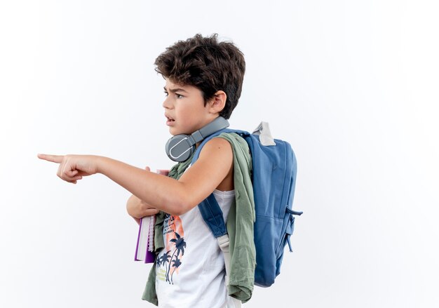 Looking at side confused little schoolboy wearing back bag and headphones points at side isolated on white background