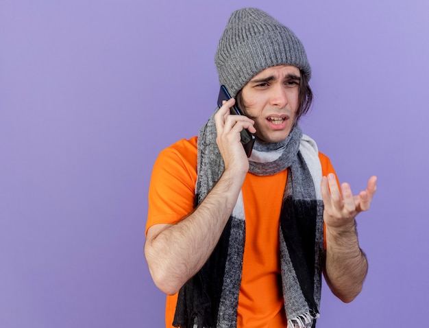 スカーフと冬の帽子をかぶって混乱している若い病気の人が紫色の背景で隔離の電話で話す