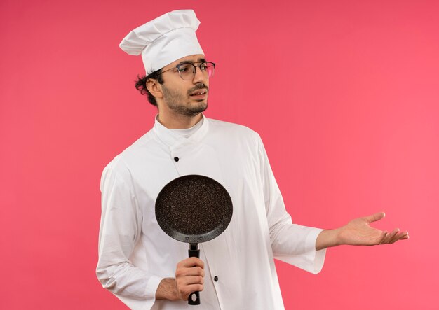 Looking at side confused young male cook wearing chef uniform and glasses holding frying pan and points with hand to side 