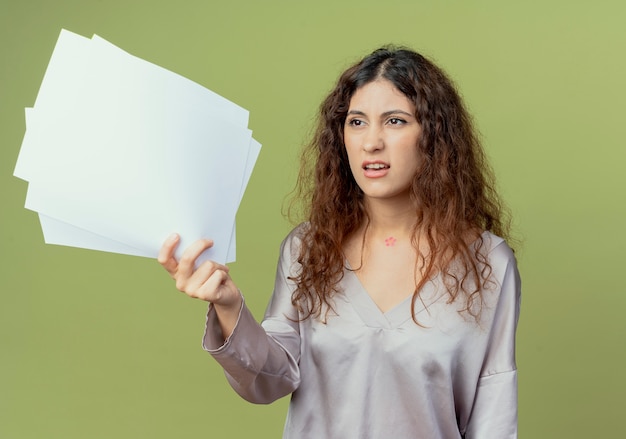 Looking at side confused young pretty female office worker holding papers isolated on olive green wall