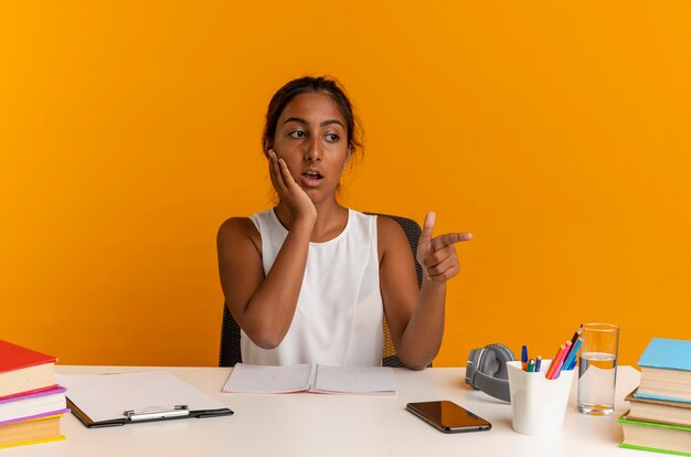 Looking at side confused young schoolgirl sitting at desk with school tools points at side and putting hand on cheek 