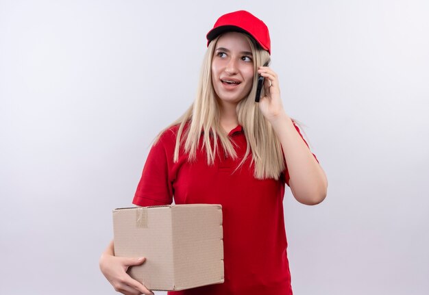 Looking at side delivery young woman wearing red t-shirt and cap in dental brace holding box and pseakes on phone on isolated white wall