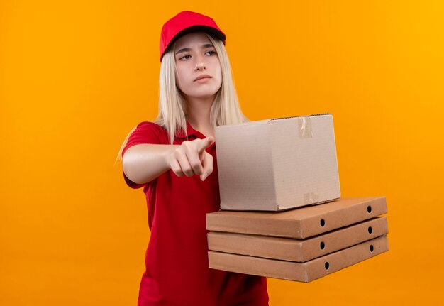 Looking at side delivery young woman wearing red t-shirt and cap holding pizza box points to side on isolated orange wall