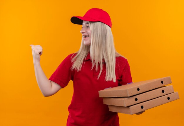 Глядя на боковую доставку, молодая женщина в красной футболке и кепке держит коробку для пиццы, показывая жест да на изолированной оранжевой стене