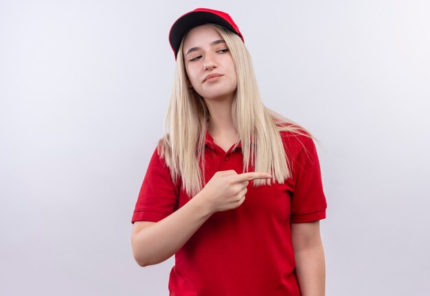 Looking at side delivery young woman wearing red t-shirt and cap points to side on isolated white wall