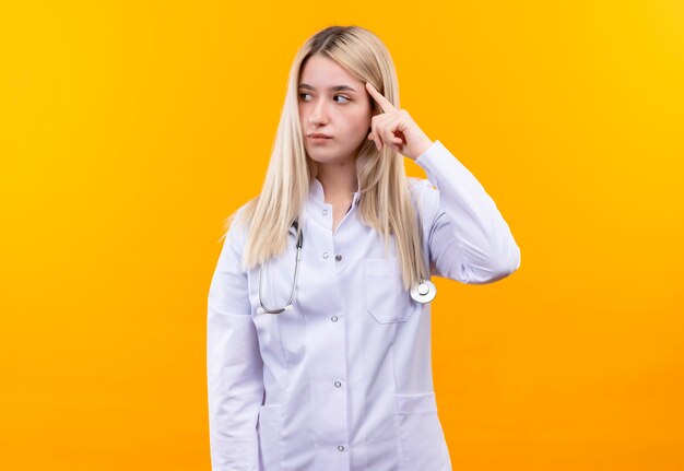 Глядя на бокового доктора, молодая девушка со стетоскопом в медицинском халате положила палец на голову на изолированной желтой стене