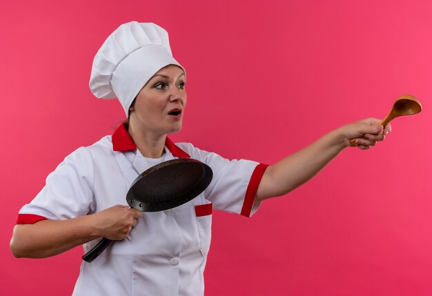 Глядя на впечатленную женщину-повара средних лет в униформе шеф-повара, держащую сковороду и протягивающую ложку в сторону на изолированной розовой стене