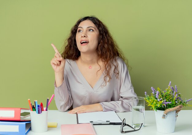 Looking at side impressed young pretty female office worker sitting at desk with office tools points at side isolated on olive background with copy space