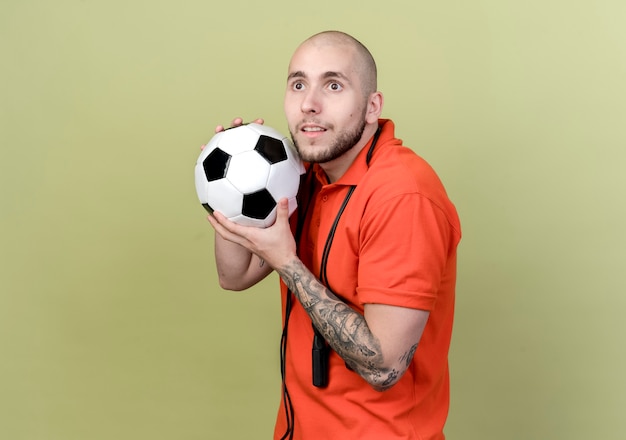 Looking at side impressed young sporty man holding ball around face with jump rope on shoulder isolated on olive green wall