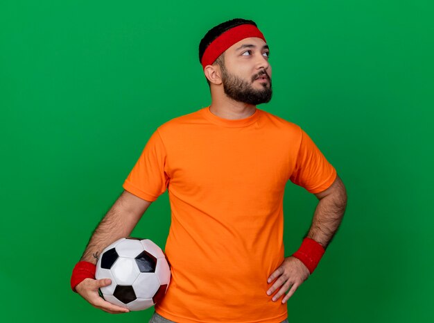 Looking at side impressed young sporty man wearing headband and wristband putting ball on hip isolated on green background
