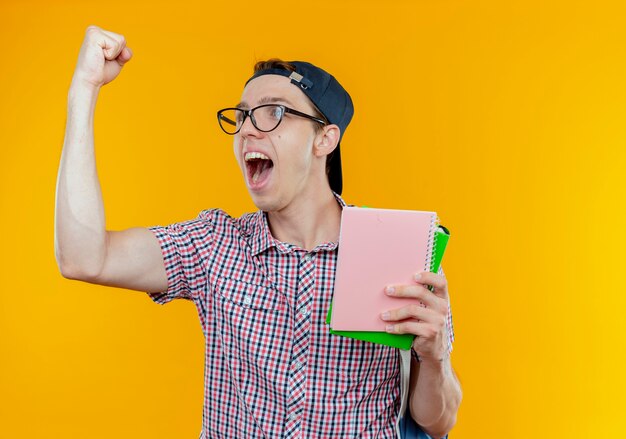 Looking at side joyful young student boy holding notebook and showing yes gesture