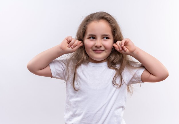 Looking at side little school girl wearing white t-shirt closed ears on isolated white wall