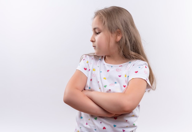 Looking at side little school girl wearing white t-shirt crossing hands on isolated white wall