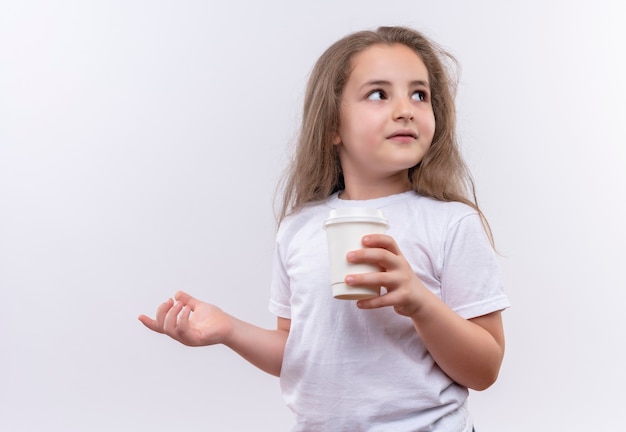 Глядя на сторону маленькой школьницы в белой футболке, держащей чашку кофе на изолированной белой стене