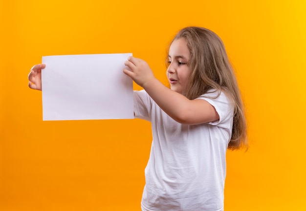 Looking at side little school girl wearing white t-shirt holding paper on isolated orange wall