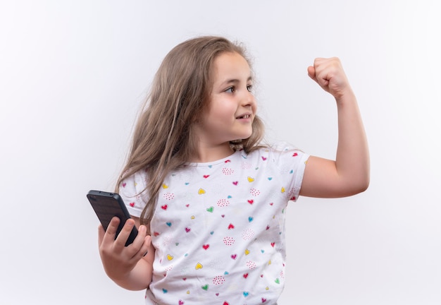 Глядя на сторону маленькой школьницы в белой футболке, держащей телефон, показывая сильный жест на изолированной белой стене