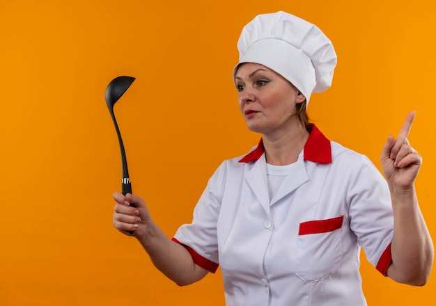 Looking at side middle-aged female cook in chef uniform holding ladle points finger to up on isolated yellow wall with copy space