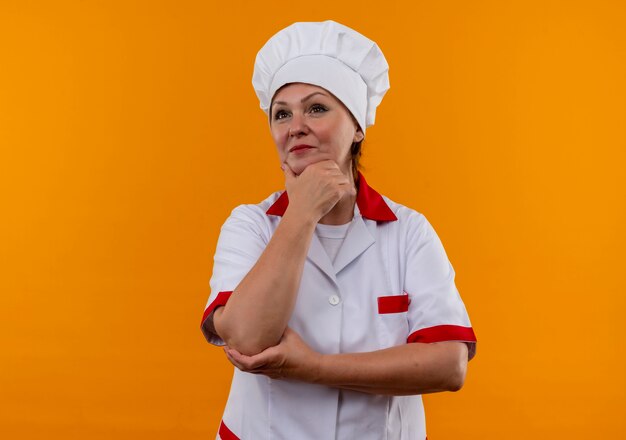 孤立した黄色の壁のあごに手を置いてシェフの制服を着た中年女性料理人の側を見て