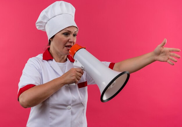 Looking at side middle-aged female cook in chef uniform speak on loudspeaker on isolated pink wall