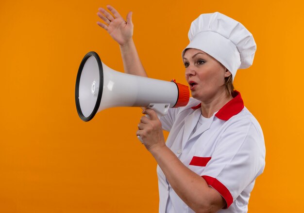 Guardando il cuoco femminile di mezza età laterale in uniforme del cuoco unico speak sull'altoparlante che solleva la mano sulla parete gialla isolata