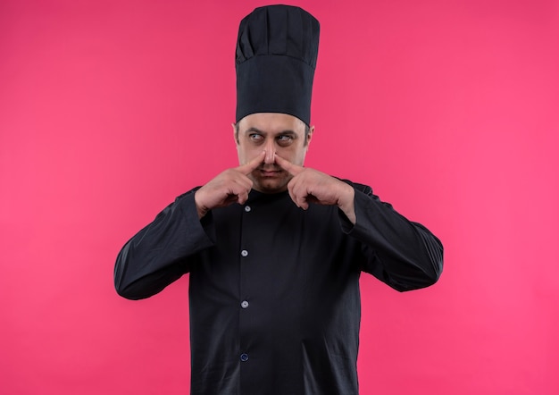 Looking at side middle-aged male cook in chef uniform closed nose on isolated pink wall