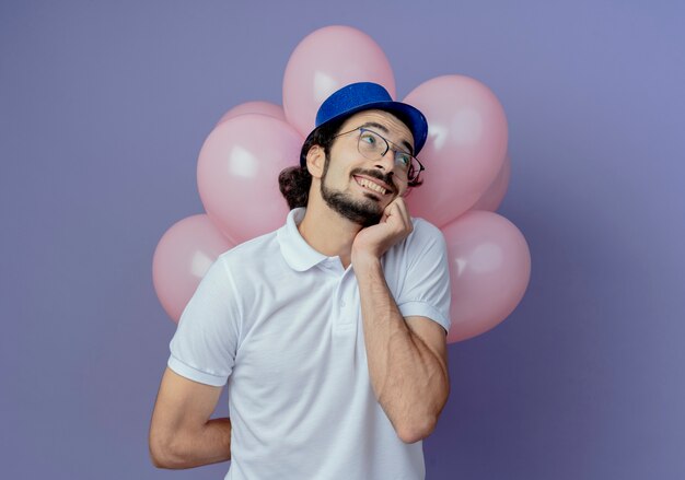 Глядя в сторону довольного красивого мужчины в очках и синей шляпе, стоящего перед воздушными шарами и положившего руку на подбородок, изолированного на фиолетовом фоне