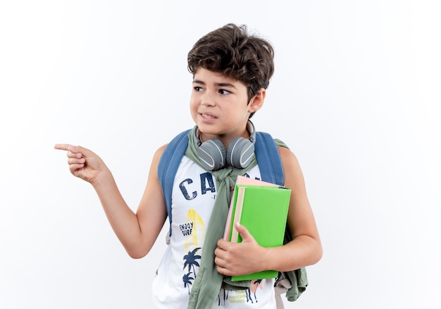 Looking at side pleased little schoolboy wearing back bag and headphones holding book and points at side isolated on white background with copy space