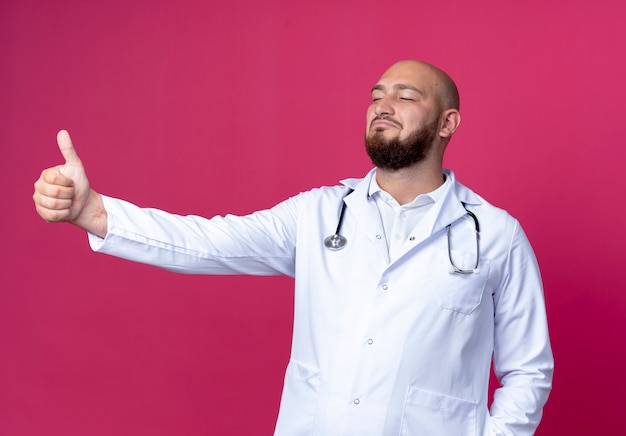 Looking at side pleased young bald male doctor wearing medical robe and stethoscope his thumb up isolated on pink background