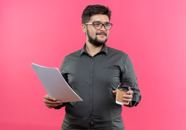 Looking at side pleased young businessman wearing glasses holding paper and cup of coffee isolated on pink background