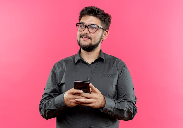 Looking at side pleased young businessman wearing glasses holding phone isolated on pink background