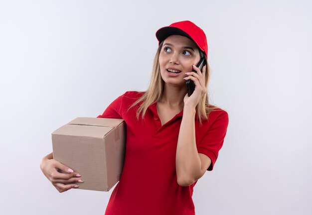 Глядя на сторону довольная молодая доставщица в красной форме и кепке, держащая коробку, и разговаривает по телефону, изолированному на белой стене