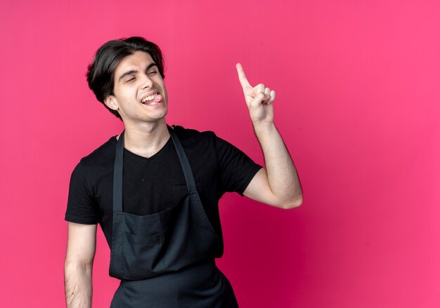 looking at side pleased young handsome male barber in uniform showing tongue and points at up