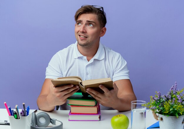 青い背景で隔離の本を保持している学校のツールと机に座って喜んで若いハンサムな男子生徒の側を見て