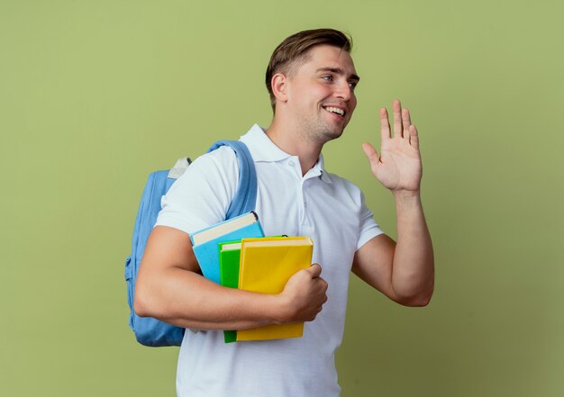 looking at side pleased young handsome male student wearing back bag holding books and raising hand isolated on olive green background