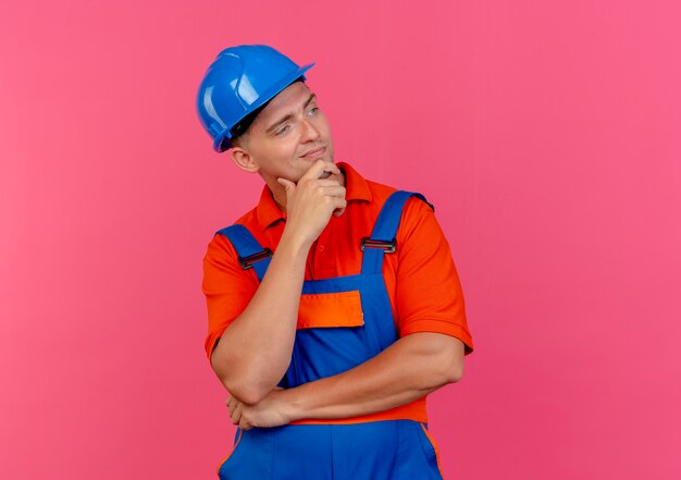 Looking at side pleased young male builder wearing uniform and safety helmet putting hand under chin 