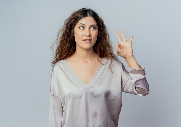 Looking at side pleased young pretty female office worker showing okey gesture isolated on white wall