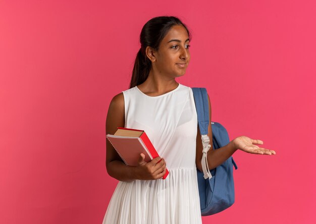 Looking at side pleased young schoolgirl wearing backpack holding book with notebook and points with hand at side  