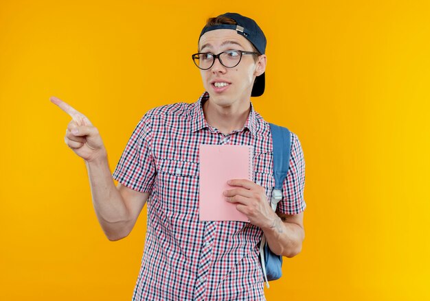 Looking at side pleased young student boy wearing backpack and glasses and cap holding notebook and points at side 