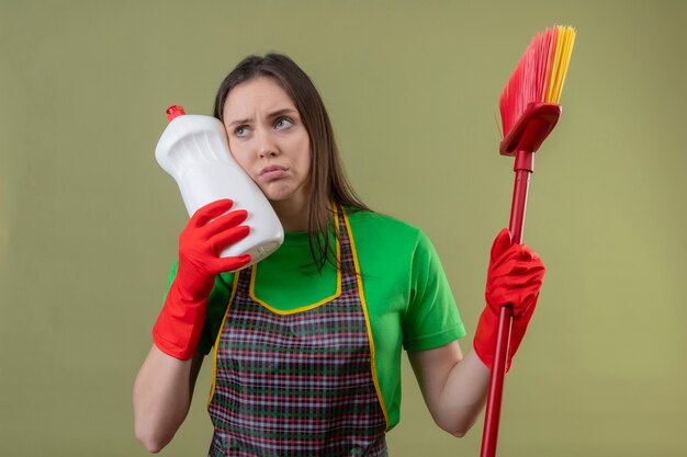 モップを保持している赤い手袋で制服を着ている悲しい掃除の若い女性の側を見て、孤立した緑の壁の頬に洗浄剤を置きます