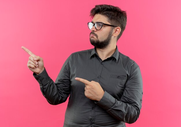 Looking at side sad young businessman wearing glasses points at side isolated on pink wall with copy space