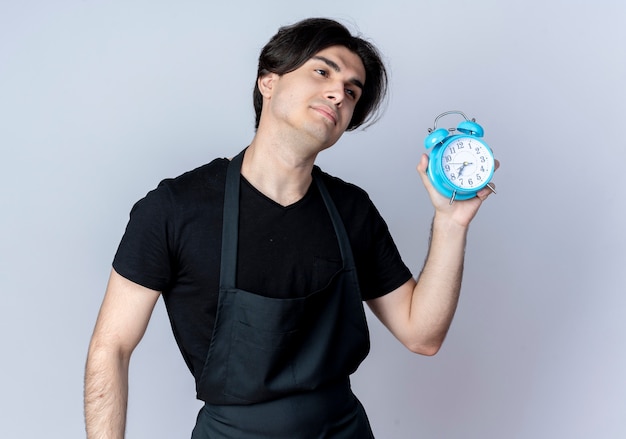 Looking at side sad young handsome male barber in uniform holding alarm clock isolated on white background