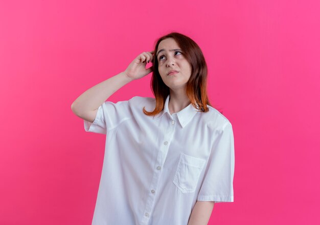 Looking at side sad young redhead girl putting hand on head isolated on pink background