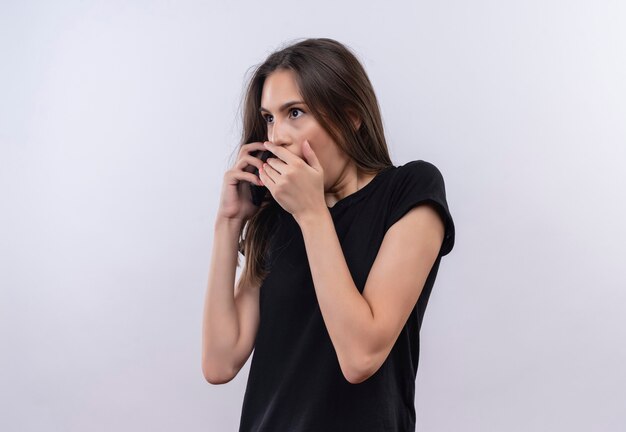 Looking at side scared young caucasian girl wearing black t-shirt put her hands around mouth on isolated white wall