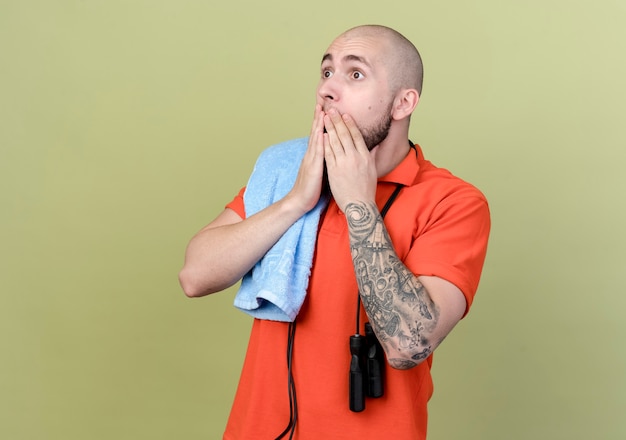 Looking at side scared young sporty man with towel and jump rope on shoulder covered mouth with hands 